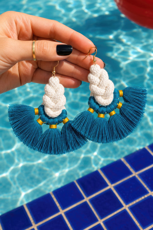A hand holding a pair of turquoise and white tassel earrings with gold accents, set against a backdrop of a blue tiled floor and a body of water.