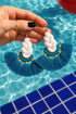 A hand holding a pair of turquoise and white tassel earrings with gold accents, set against a backdrop of a blue tiled floor and a body of water.