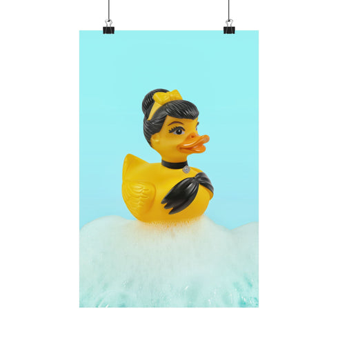 A chic rubber duck styled like Audrey Hepburn’s Holly Golightly, wearing a high bun, yellow bow, black gloves, and a sparkly choker. The duck floats in a pile of foam and is framed in white on a bubbly bathroom shelf with a red tile backdrop and soap dispenser.