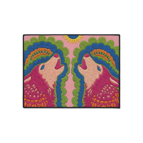 A rectangular outdoor doormat featuring a symmetrical graphic illustration of two howling wolves facing upward in profile. The design utilizes a vibrant psychedelic folk art style with a color palette of magenta pink wolves, peach muzzles, and scalloped layers of lime green and cyan blue foliage. A marigold yellow sun sits at the top center. The background is a dusty rose pink. The texture is a smooth, durable flat 2D print on polyester with a black non-slip rubber border.