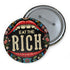 A round pin-back button with a bold, embroidered-style design featuring the phrase "EAT THE RICH" in large, vintage-style lettering. The typography is framed by a set of open red lips with detailed stitching, revealing white teeth. Surrounding the mouth are intricate floral patterns in shades of blue, red, and green, adding a decorative, folk-art-inspired touch. The button has a black background, making the details and lettering stand out