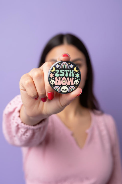 A round pinback button featuring a solid black background covered in a retro pastel colored design. The center reads 25TH NOW in a groovy bubbly font using pale pink and mint green referencing the 25th amendment. Surrounding the text is a maximalist cluster of cute but edgy motifs including smiling white skulls, pastel rainbows, pale yellow crescent moons, daisies, and a pink and white spotted mushroom. The button has a glossy laminate finish and a secure metal safety pin backing.