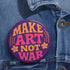 A round, glossy purple pinback button with the words "MAKE ART NOT WAR" in a groovy, retro 70s-style font. The word "MAKE" is in orange, "ART" is in a vibrant pink, and "NOT WAR" is in orange. The text is surrounded by small, stylized flower and star details. This psychedelic protest pin is a piece of activist merch with a strong metal back and safety pin clasp, perfect as an anti-war or social justice accessory for a jean jacket or backpack. The pin has a handmade, indie aesthetic.