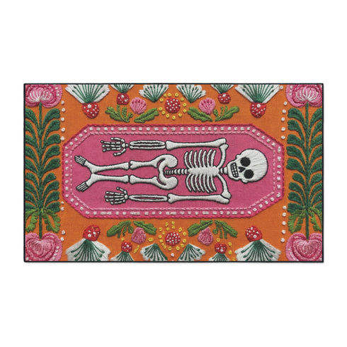 A rectangular terracotta orange doormat featuring a central illustration of a white skeleton lying inside a pink coffin shape. The design is surrounded by a symmetrical pattern of green leaves, pink flowers, and red and white spotted mushrooms. The imagery uses a trompe-l'œil effect to mimic the texture of raised needlepoint embroidery, but the surface is flat printed polyester. The mat is framed with a solid black edge.