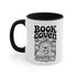 A two-tone ceramic mug with a glossy white exterior and a contrasting black interior, handle, and rim. The front of the mug features a black, rectangular graphic with the words "BOOK COVEN" in a bold, wavy, 70s-inspired retro font. Below the text is an occult-style illustration of an open book on a stand. An all-seeing eye with a snake coiled around it emerges from the book's pages, radiating lines of light. Smaller books float around the central image, completing the witchy, bookish design