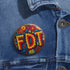 Round pinback metal button featuring an embroidered-style design on a vibrant, multicolored background. The bold, textured letters "FDT" are prominently displayed in red with intricate golden-yellow stitching details, outlined in dark blue. Surrounding the text is an ornate floral pattern with embroidered-style flowers in shades of red, orange, blue, and yellow, densely arranged for a rich, decorative effect. The outer edge is bordered with a zigzag stitching effect in red and blue. The button's back is sil