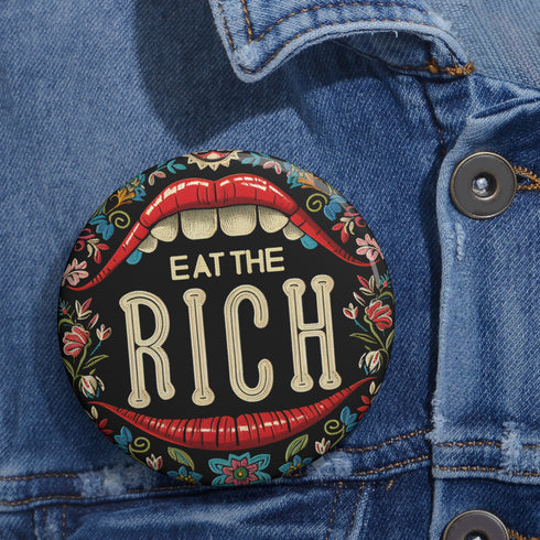A round pin-back button with a bold, embroidered-style design featuring the phrase "EAT THE RICH" in large, vintage-style lettering. The typography is framed by a set of open red lips with detailed stitching, revealing white teeth. Surrounding the mouth are intricate floral patterns in shades of blue, red, and green, adding a decorative, folk-art-inspired touch. The button has a black background, making the details and lettering stand out