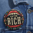 A round pin-back button with a bold, embroidered-style design featuring the phrase "EAT THE RICH" in large, vintage-style lettering. The typography is framed by a set of open red lips with detailed stitching, revealing white teeth. Surrounding the mouth are intricate floral patterns in shades of blue, red, and green, adding a decorative, folk-art-inspired touch. The button has a black background, making the details and lettering stand out