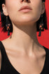 A close-up of a woman's face wearing black dangling earrings against a red background.