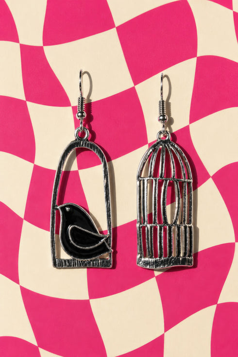 two silver earrings with bird designs, one in a cage and one without a cage, set against a pink and white checkered background.