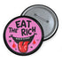 Round pink pinback button featuring a cartoon-style open mouth with sharp teeth, a red tongue, and dripping red details. The bold black text reads “EAT THE RICH” in a hand-drawn style. Glossy black rim with a vibrant, rebellious graphic design.