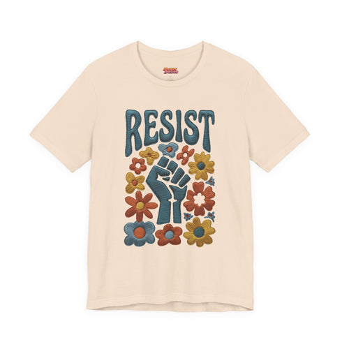 Cream unisex t-shirt featuring a centered design with dark teal raised fist surrounded by folk art botanical elements: crimson and red 5-petal daisies, golden yellow and orange flowers, cornflower blue petals, and small scattered wildflower accents arranged in a symmetrical medallion composition. Dark teal "RESIST" text appears above the fist. The design uses faux embroidery 2D print with dimensional texture detail. Shown on unisex fit with ribbed knit collar and short sleeves.