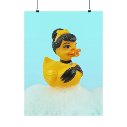 A chic rubber duck styled like Audrey Hepburn’s Holly Golightly, wearing a high bun, yellow bow, black gloves, and a sparkly choker. The duck floats in a pile of foam and is framed in white on a bubbly bathroom shelf with a red tile backdrop and soap dispenser.