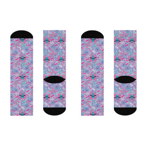 Knee-high socks worn on legs, featuring a pastel blue background with a quirky pattern. The pattern includes pink and teal skulls, some adorned with bows, alongside images of cherries, hearts, and additional bow designs. The socks have black toes and heels, contrasting with the colorful pattern. The overall effect is a playful mix of cute and edgy elements.