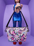 Small vegan leather crossbody purse with a real pebble grain texture and adjustable black strap. The flat printed artwork features a scattered all over pattern of pink skulls, purple and pastel blue winged bats, pink fanged mouths, and loose eyeballs set against a cream colored background. The bag is shown with black and silver hardware including a black top zipper, black zipper pull, black strap tabs, and silver metal rings and clips. The compact rectangular bag measures 8.5 by 7 by 2 inches.