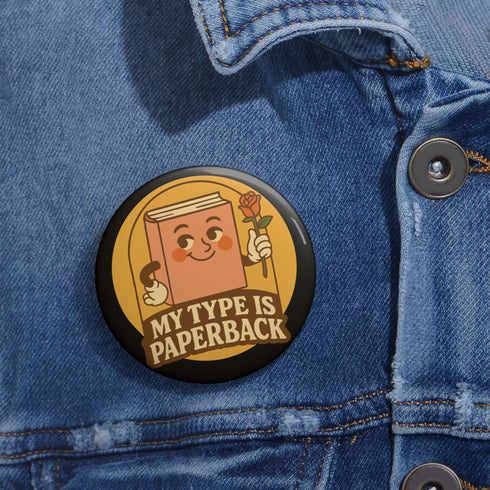 A glossy black round pin-back button with a retro 1970s-style design. The center shows a smiling cartoon paperback book holding a red rose, colored light brown with white gloves and feet. Below, “MY TYPE IS PAPERBACK” appears in chunky brown-and-tan 70s lettering. This bookish flair piece has a high-gloss, scratch-resistant mylar finish, secure safety pin backing, and comes in two- or three-inch sizes.