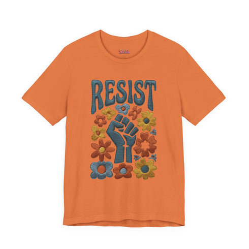 Cream unisex t-shirt featuring a centered design with dark teal raised fist surrounded by folk art botanical elements: crimson and red 5-petal daisies, golden yellow and orange flowers, cornflower blue petals, and small scattered wildflower accents arranged in a symmetrical medallion composition. Dark teal "RESIST" text appears above the fist. The design uses faux embroidery 2D print with dimensional texture detail. Shown on unisex fit with ribbed knit collar and short sleeves.
