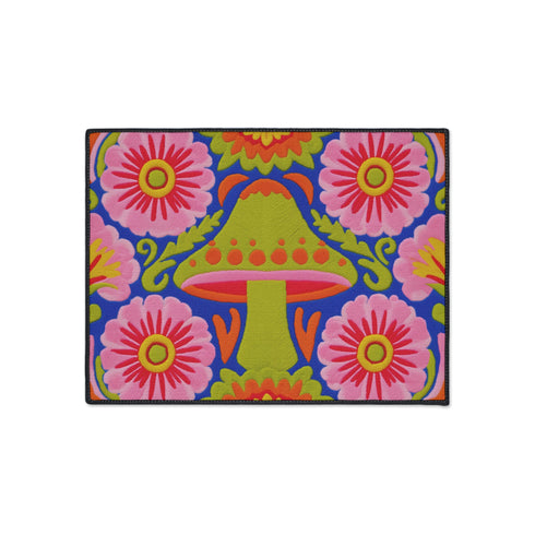 A rectangular polyester mat featuring a large central mushroom with a lime green cap, orange spots, and a pink underside. The design is surrounded by four large magenta and pink radial flowers with yellow centers. Bright green leaves and orange scrolling vines fill the spaces on a deep cobalt blue background. The artwork uses a tapestry-style visual effect to mimic the look of raised embroidery on a flat, printed surface. The mat is finished with a clean black stitched edge.