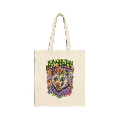 Black or natural cotton canvas tote bag with “The Future is Femme & Feral” design featuring a rainbow-colored snarling opossum with flowers and feminist symbols, printed on both sides with long matching canvas handles.