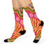 A pair of vibrant crew socks featuring a maximalist 1970s inspired collage pattern. The design includes blocks of hot pink with white daisy flowers and yellow centers, clashing playfully with panels of mustard yellow and orange tiger stripes. Wavy psychedelic lines in grass green and orange frame the top cuff. The socks have a solid black heel and toe cap. The fabric is a smooth, stretchy polyester blend with a flat knit texture and bright colors.