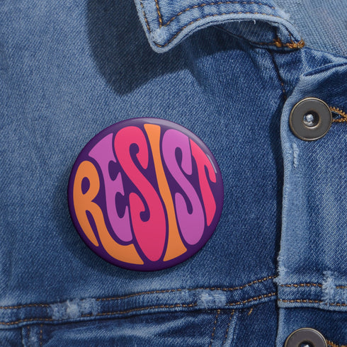 Close-up of a glossy, round purple pinback button. The button features the word "RESIST" in a bold, 70s-style psychedelic font that fills the entire surface. The bubbly, wavy letters alternate in color between a vibrant orange, a light peach-pink, and a deep magenta, creating a powerful and retro statement piece. The design has a distinct groovy and feminist protest aesthetic, making it a perfect accessory for jackets, bags, or activist apparel.