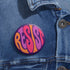 Close-up of a glossy, round purple pinback button. The button features the word "RESIST" in a bold, 70s-style psychedelic font that fills the entire surface. The bubbly, wavy letters alternate in color between a vibrant orange, a light peach-pink, and a deep magenta, creating a powerful and retro statement piece. The design has a distinct groovy and feminist protest aesthetic, making it a perfect accessory for jackets, bags, or activist apparel.