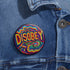 Round black pinback button with neon embroidery-style waves surrounding bold yellow and red "DISOBEY" text.