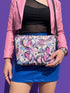 Small vegan leather crossbody purse with a real pebble grain texture and adjustable black strap. The flat printed artwork features a scattered all over pattern of pink skulls, purple and pastel blue winged bats, pink fanged mouths, and loose eyeballs set against a cream colored background. The bag is shown with black and silver hardware including a black top zipper, black zipper pull, black strap tabs, and silver metal rings and clips. The compact rectangular bag measures 8.5 by 7 by 2 inches.