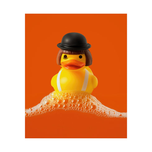 A fine art print of a rubber duck dressed like Alex from A Clockwork Orange, featuring a black bowler hat, blunt bob, and white suspenders. The duck floats in orange-tinged bubbles and is framed in white on a glossy pink tile bathroom shelf.