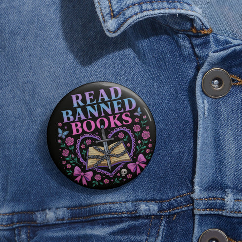 Glossy black pinback button that says “Read Banned Books” in purple and teal letters, featuring a chained book, roses, bows, and floral accents, styled on colorful books and pastel backgrounds.