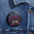 Glossy black pinback button that says “Read Banned Books” in purple and teal letters, featuring a chained book, roses, bows, and floral accents, styled on colorful books and pastel backgrounds.