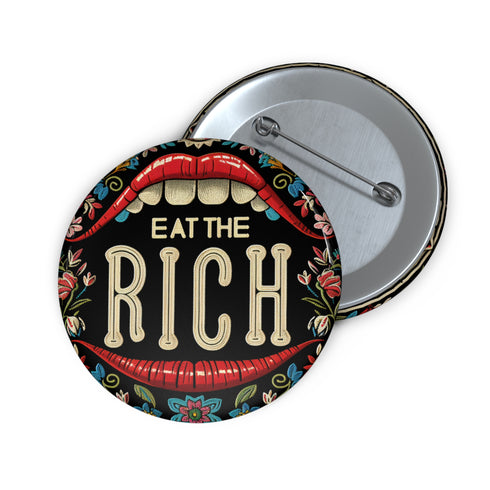 A round pin-back button with a bold, embroidered-style design featuring the phrase "EAT THE RICH" in large, vintage-style lettering. The typography is framed by a set of open red lips with detailed stitching, revealing white teeth. Surrounding the mouth are intricate floral patterns in shades of blue, red, and green, adding a decorative, folk-art-inspired touch. The button has a black background, making the details and lettering stand out
