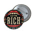 A round pin-back button with a bold, embroidered-style design featuring the phrase "EAT THE RICH" in large, vintage-style lettering. The typography is framed by a set of open red lips with detailed stitching, revealing white teeth. Surrounding the mouth are intricate floral patterns in shades of blue, red, and green, adding a decorative, folk-art-inspired touch. The button has a black background, making the details and lettering stand out