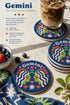 A close-up photograph of a round, glossy ceramic coaster featuring a bold retro pop art design. The background is a deep royal blue. In the center are two symmetrical profile faces of twins looking at each other, featuring bright green skin, closed eyes, and red cheeks. They have wavy white and pale pink hair with red accents. The word Gemini is arched at the top in a retro white font. The design is decorated with a green Gemini symbol, pink and red hearts, and white starbursts.