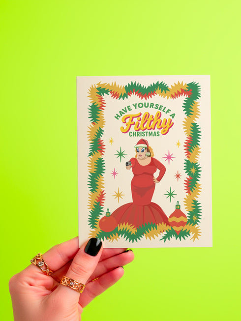A 5x7 pearlescent holiday card by HoneyDazed. The card illustration, inspired by John Waters, features the drag queen Divine in a red dress with a Santa hat. She points a gun, withtext reads "Have Yourself a Filthy Christmas" in campy font. The card is bordered by garland. Iinside of the card reads: "Season's Sleazings" in red and green text. This kitschy greeting card is a perfect piece of cult film memorabilia and a funny adult humor gift for fans of queer art.