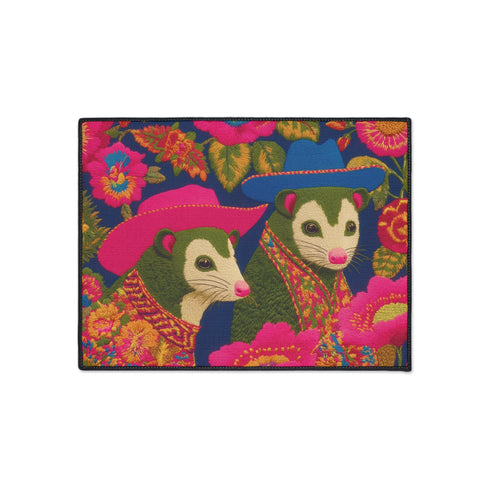 A rectangular non-slip doormat with a black trimmed edge. The mat features a vibrant, colorful folk art illustration on a dark navy blue background with a faux embroidered texture. The design depicts two friendly opossums, rendered in an olive green color with white faces and pink noses. They are surrounded by large, stylized flowers and leaves in shades of hot pink, red, turquoise, and yellow, creating a busy, maximalist pattern. The overall aesthetic is eclectic, whimsical, & highly detailed.