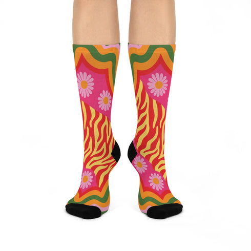 A pair of vibrant crew socks featuring a maximalist 1970s inspired collage pattern. The design includes blocks of hot pink with white daisy flowers and yellow centers, clashing playfully with panels of mustard yellow and orange tiger stripes. Wavy psychedelic lines in grass green and orange frame the top cuff. The socks have a solid black heel and toe cap. The fabric is a smooth, stretchy polyester blend with a flat knit texture and bright colors.