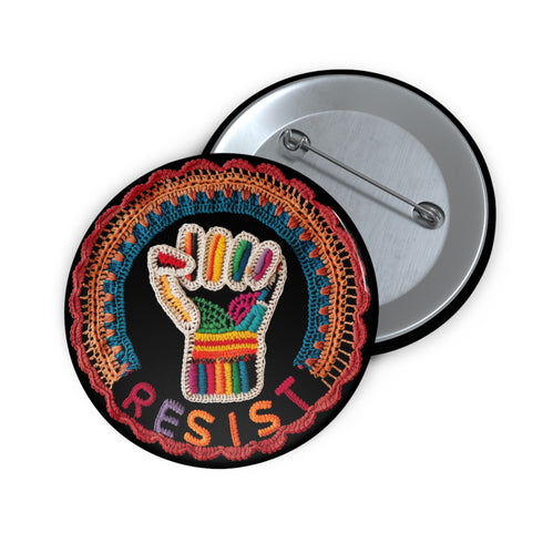 Round pinback metal button with an embroidered-style design on a black background. The center features a raised fist symbol stitched in a multicolored pattern, incorporating rainbow hues and folk-inspired textures. The fist is encircled by a vibrant, lace-like border in shades of blue, orange, and red, resembling intricate embroidery. Below the fist, the word "RESIST" is spelled out in bold, stitched-textured letters in red and orange. The button has a silver metal back with a secure pin closure.