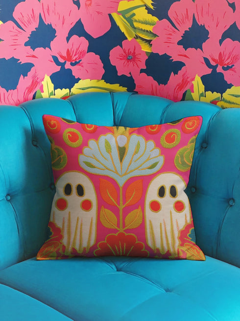 A vibrant, colorful throw pillow with a floral design sits on a blue tufted chair, with a bold, colorful floral wallpaper in the background.