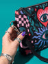 Small vegan leather crossbody purse with a real pebble grain texture and adjustable black strap. The flat printed artwork features a central blue sunburst evil eye motif below a coral pink sacred heart eye, surrounded by pink crescent moons, multicolored stars, flowing teal botanical vines, and wavy lavender and black checkerboard distortion banners. Set against a dark background with silver metal hardware including a top zipper, zipper pull, and strap clips. Measures 8.5 by 7 by 2 inches.