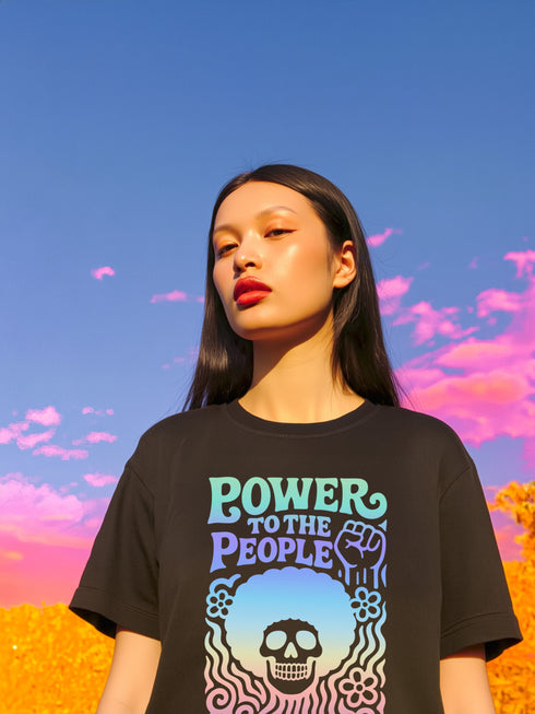 A detailed view of a black unisex graphic t-shirt made of soft cotton, shown flat. The shirt features a large, rectangular design on the chest inspired by 70s psychedelic protest art. The words "POWER TO THE PEOPLE" are printed at the top in a thick, rounded, groovy font. Below the text is a stylized human skull with a large, perfectly round afro hairstyle. The entire design, including the text, skull, and afro, is filled with a vibrant color gradient that shifts from deep purple to hot pink.