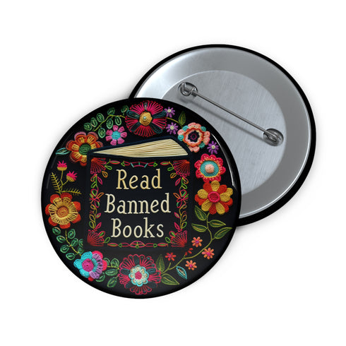Round black pinback button with "Read Banned Books" in gold lettering on an open book image, surrounded by vibrant embroidery-style flowers in pink, orange, blue and green. Folk art-inspired literary protest pin with glossy finish and safety pin backing, available in 2" and 3" sizes.