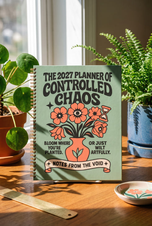 A sage green hardcover spiral planner for 2027-2028 with the title "The 2027 Planner of Controlled Chaos" in a funky, retro 70s-style font. The cover features a groovy illustration of orange and pink flowers in a vase, with one flower depicted as an eyeball. The text reads "Bloom where you're planted. Or just wilt artfully." and "Notes from the Void." The planner has a gold metal wire-o binding. The back cover shows a decorative floral frame with a "Property Of" section