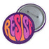 Close-up of a glossy, round purple pinback button. The button features the word "RESIST" in a bold, 70s-style psychedelic font that fills the entire surface. The bubbly, wavy letters alternate in color between a vibrant orange, a light peach-pink, and a deep magenta, creating a powerful and retro statement piece. The design has a distinct groovy and feminist protest aesthetic, making it a perfect accessory for jackets, bags, or activist apparel.