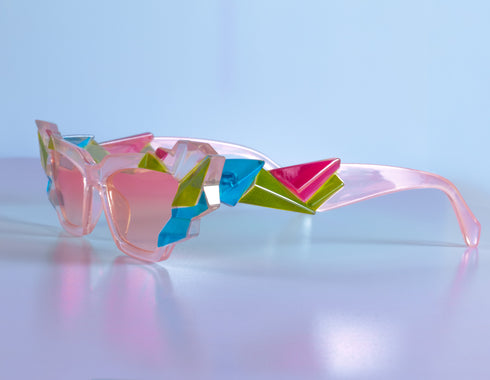 A pair of geometric sunglasses with angular frames featuring vibrant green, blue, and pink accents. The lenses are tinted light pink, adding a playful, modern twist to the bold, colorful design. These sunglasses make a striking, artistic statement
