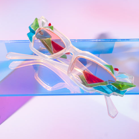 A pair of geometric sunglasses with angular frames featuring vibrant green, blue, and pink accents. The lenses are tinted light pink, adding a playful, modern twist to the bold, colorful design. These sunglasses make a striking, artistic statement