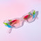 A pair of geometric sunglasses with angular frames featuring vibrant green, blue, and pink accents. The lenses are tinted light pink, adding a playful, modern twist to the bold, colorful design. These sunglasses make a striking, artistic statement