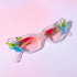 A pair of geometric sunglasses with angular frames featuring vibrant green, blue, and pink accents. The lenses are tinted light pink, adding a playful, modern twist to the bold, colorful design. These sunglasses make a striking, artistic statement
