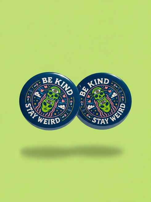 A round 1.75 inch pinback button featuring a retro psychedelic design on a dark navy blue background. The text reads BE KIND STAY WEIRD in vintage cream lettering around the border. The center illustration depicts a neon green skull stacked on top of a neon green alien head. Both the skull and alien have opening third eyes on their foreheads. Pink wavy lines frame the faces. The background is decorated with small colorful mushrooms, hearts, stars, and clouds. Glossy finish.