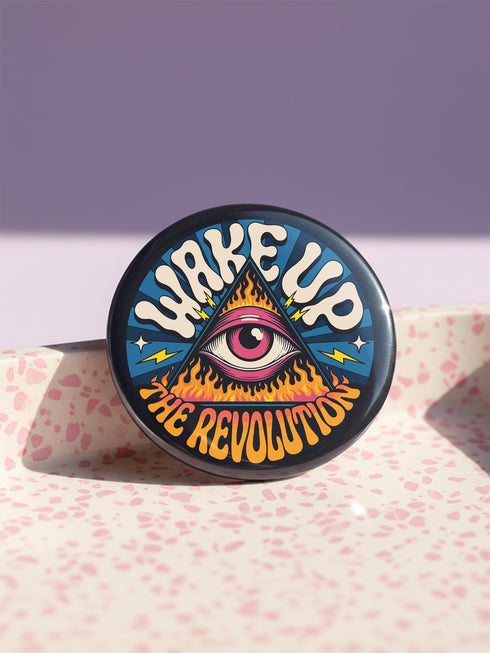 A round pinback button featuring a retro psychedelic design. The central focus is a stylized Eye of Providence in a pink triangle, surrounded by vibrant orange and yellow flames. Behind the triangle are teal and petrol blue sunburst rays. The text "WAKE UP" curves across the top in cream wavy letters, and "THE REVOLUTION" curves along the bottom in orange. The button has a glossy finish and is available in 1.75 inch or 2.25 inch sizes.